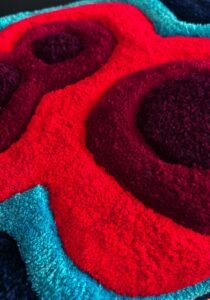 Minkšto pufo detalė iš viršaus. Iš spalvotų siūlų suformuoti raudoni ratilai, apvesti žydra ir mėlyna juostomis.